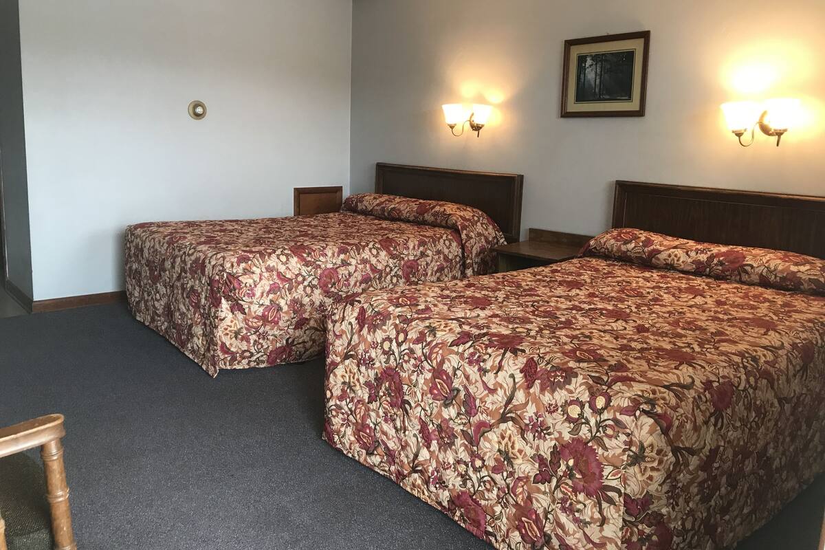 A hotel room with two single beds, a wooden headboard, a small table, and two wall-mounted lamps.