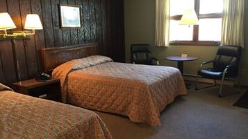 A hotel room with two beds, a wooden headboard, a small table, a chair, and a window with curtains.