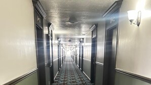 Corredor de hotel comprido e estreito, com carpete estampado, paredes claras e portas em ambos os lados.