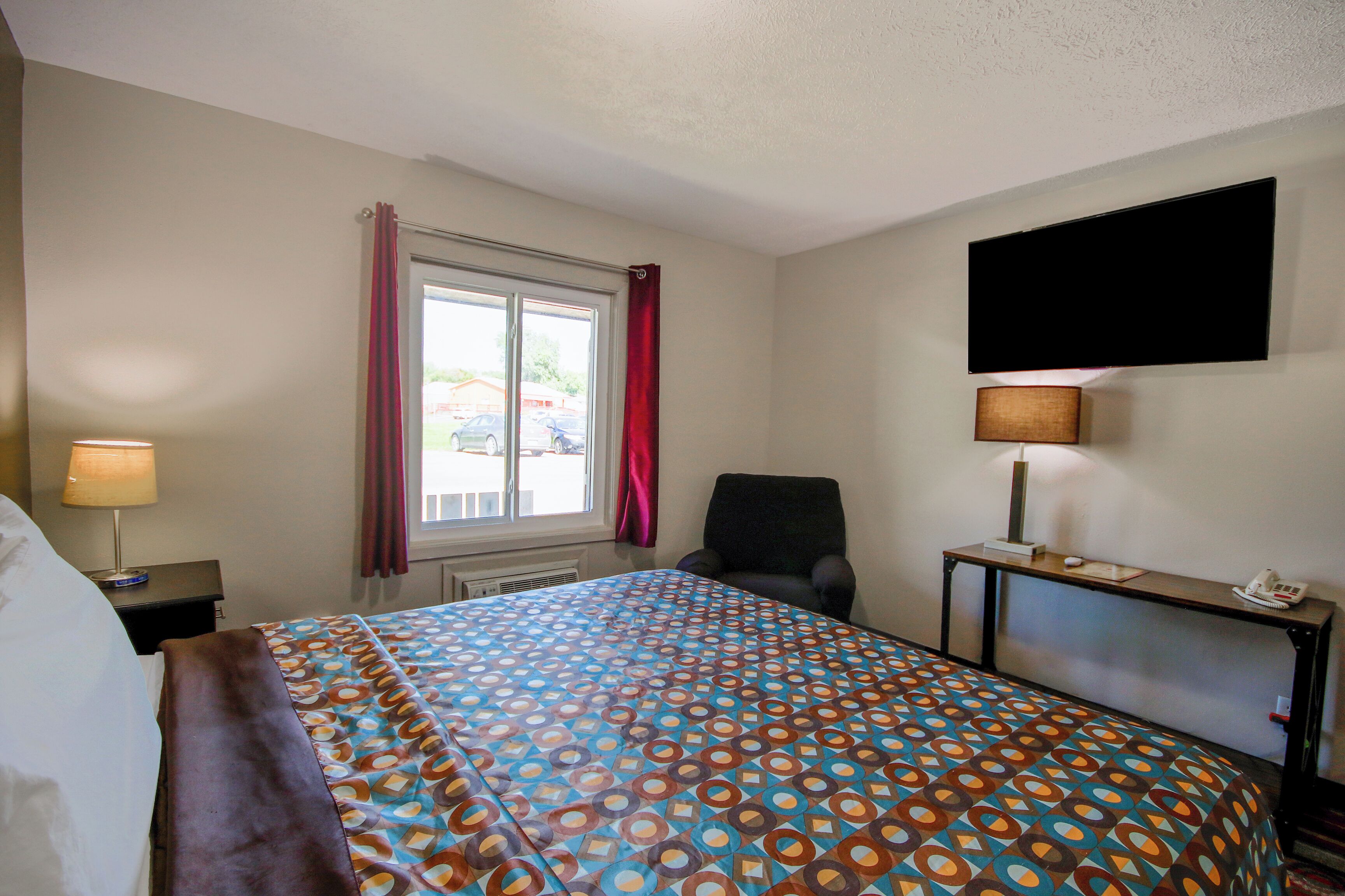 A hotel room with a bed, a chair, a desk with a lamp, a flat-screen TV, and a window with curtains.