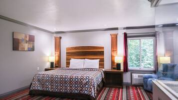 Habitación de hotel con cama de cabecera de madera, colcha estampada, alfombra a rayas negra y roja, ventana con vistas a vegetación y una pequeña zona de estar con una silla azul.