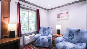 Um quarto com uma janela grande, duas poltronas com padrão de folhas, uma mesinha de madeira, uma pequena mesa com abajur e uma obra de arte na parede.