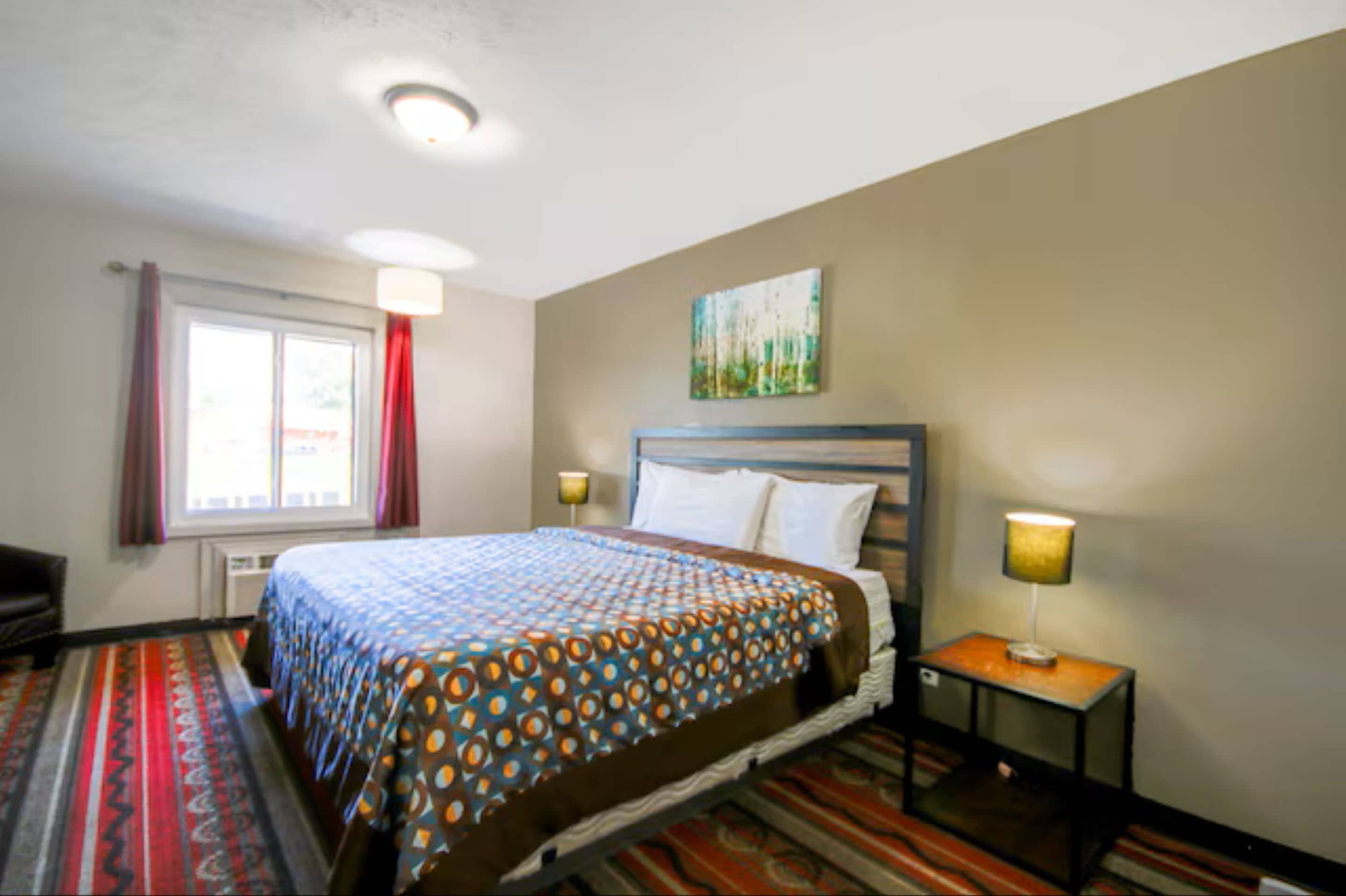 A hotel room with a bed, a nightstand, a lamp, a window with curtains, and a painting on the wall.