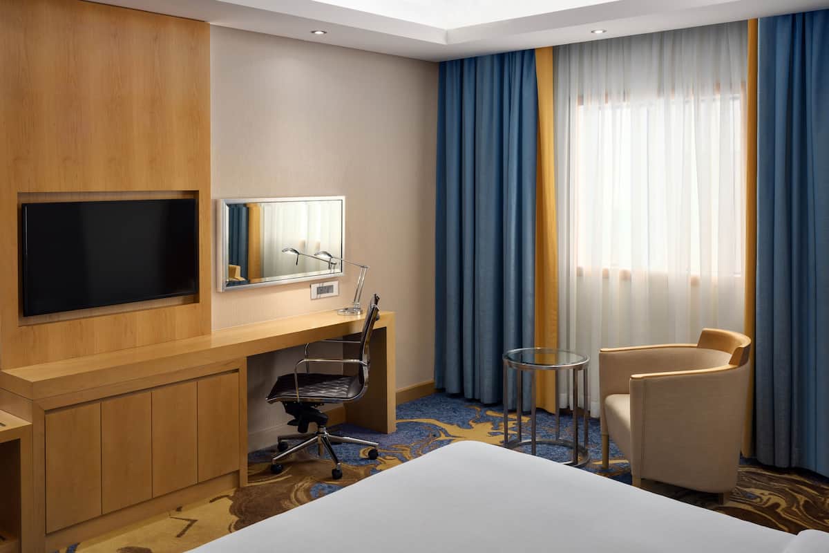 Habitación de hotel con cama, escritorio con silla, televisor, espejo y ventana con cortinas.