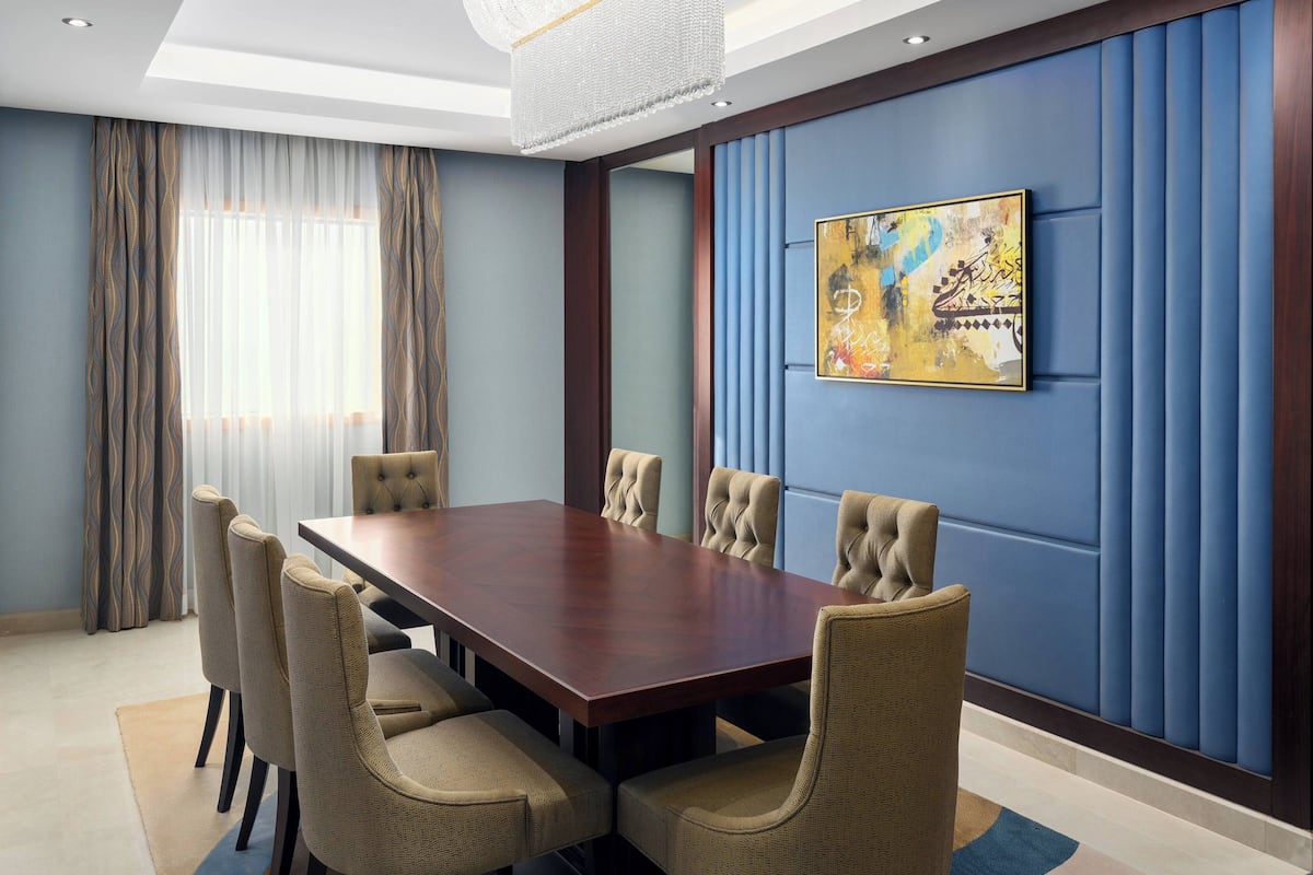 Un salón de conferencias con una mesa de madera, seis sillas tapizadas, un cuadro abstracto de gran tamaño y paredes acolchadas de color azul.