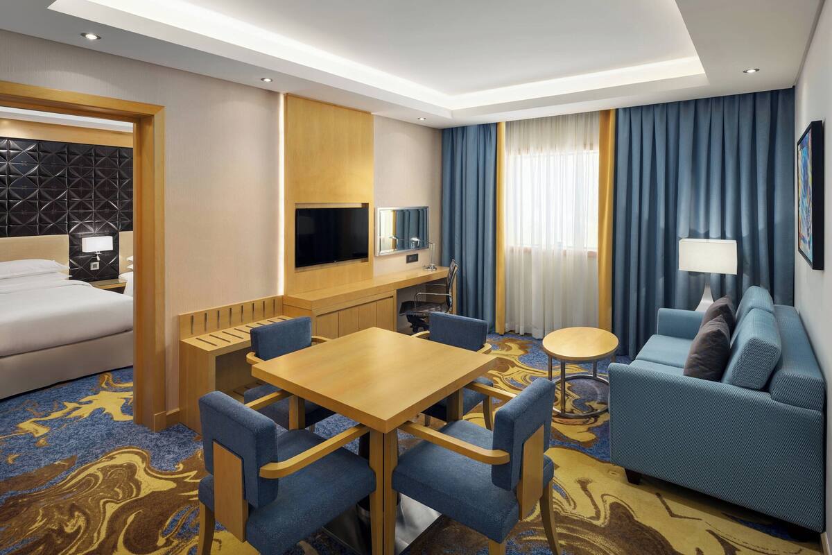 Habitación de hotel con cama, escritorio con televisor, sofá y mesa de comedor con sillas.