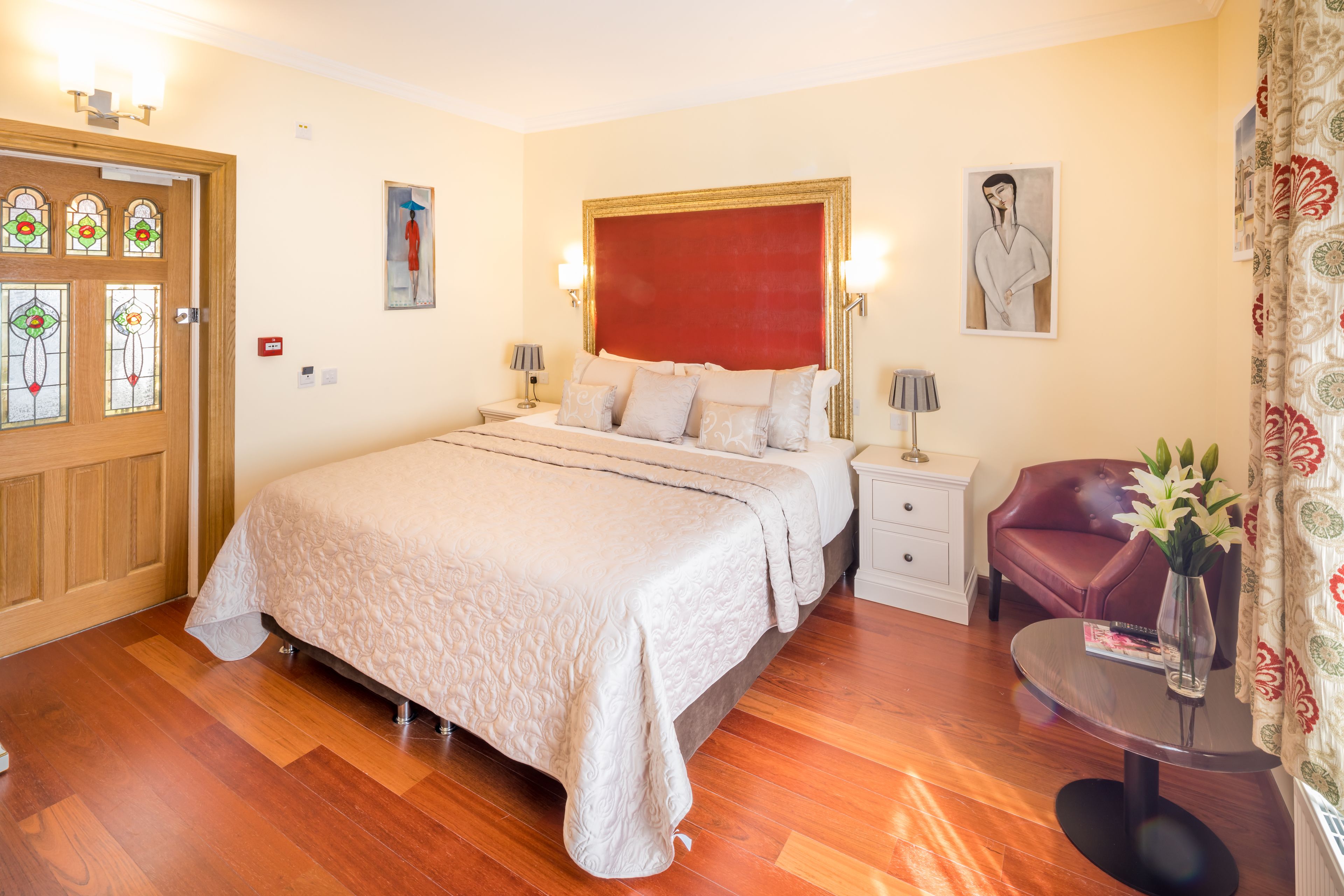 Una habitación de hotel con una cama grande, una mesita de noche, una silla, una pequeña mesa con un jarrón y una puerta con vidrieras.