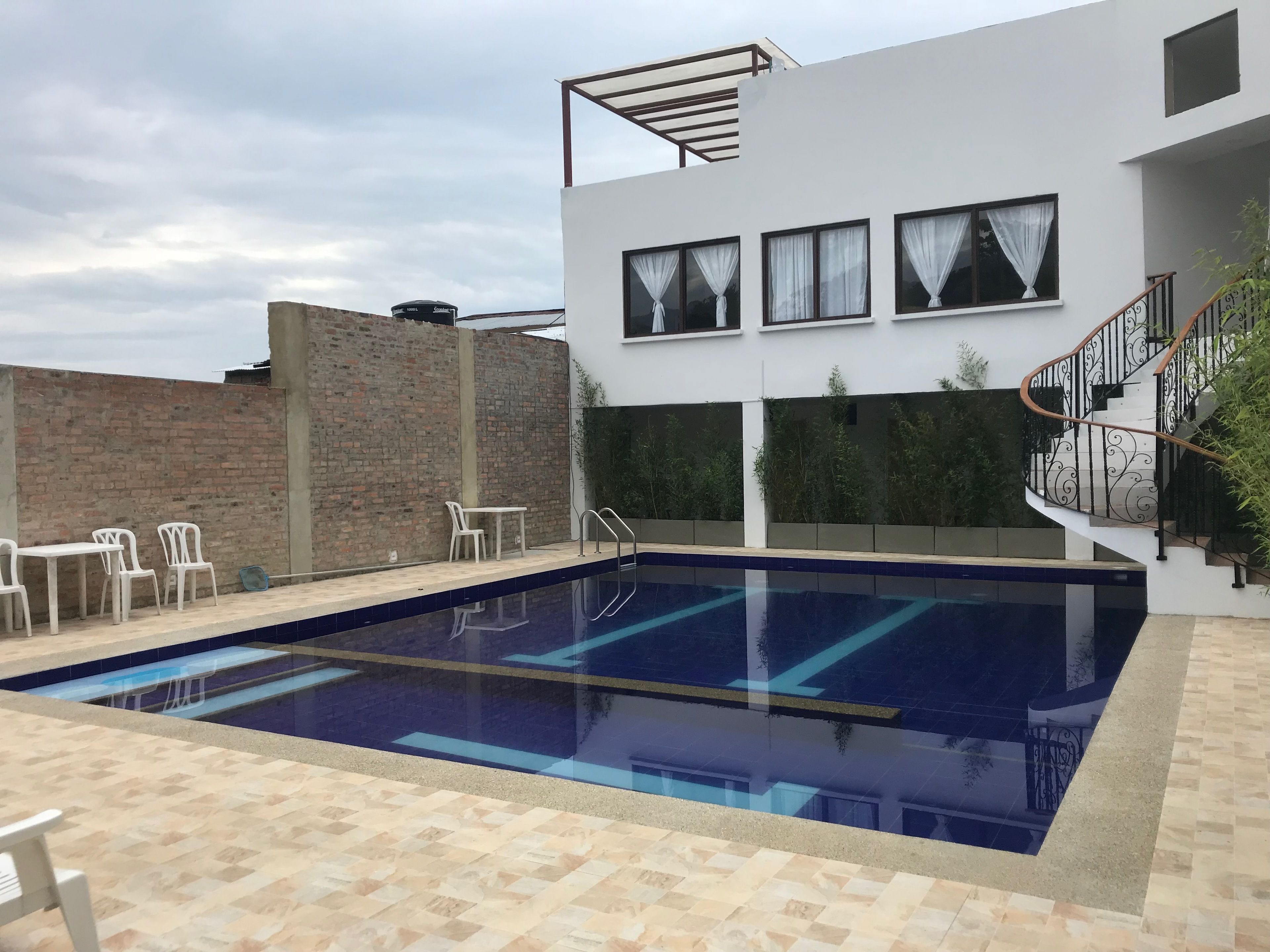 Un área de piscina moderna al aire libre con una piscina azul cristalina, un muro de ladrillo y un edificio blanco con una escalera.