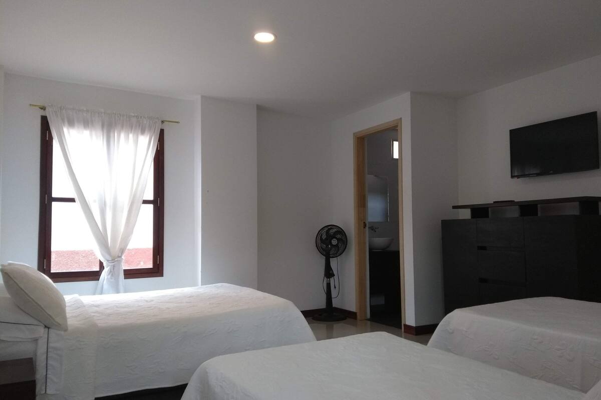 Camera d'albergo con due letti, una televisione a parete, una ventola e una finestra con tende.