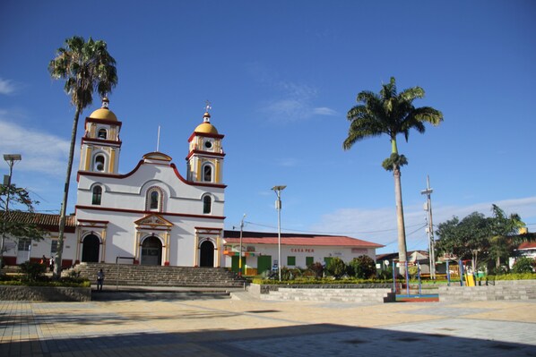 A church with two domes and a bell tower, surrounded by palm trees and a paved plaza.