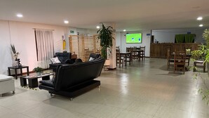 Uma sala de estar moderna com um sofá preto, uma mesa de centro e uma TV exibindo um jogo esportivo. Há uma área de jantar com cadeiras de madeira e uma mesa.