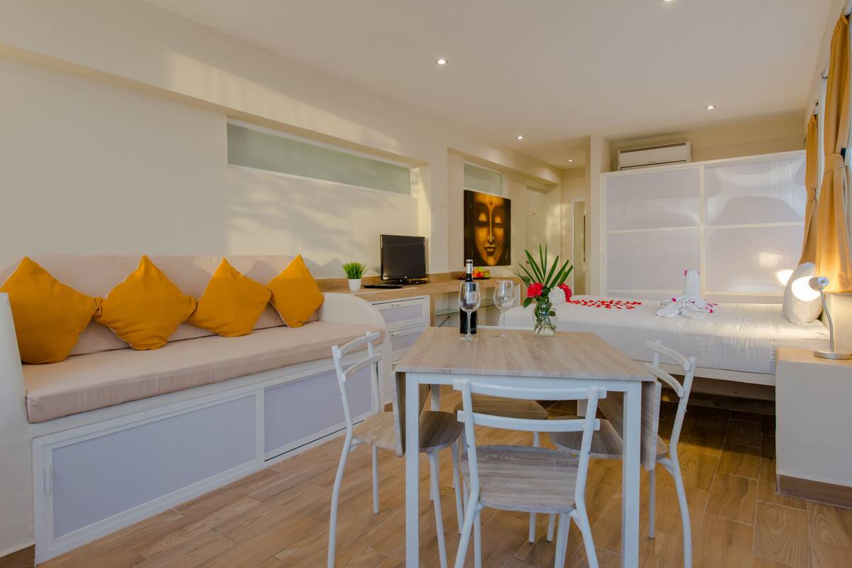 Una sala de estar moderna con un sofá, una mesa de comedor y sillas. Hay un televisor, una planta y un cuadro en la pared.