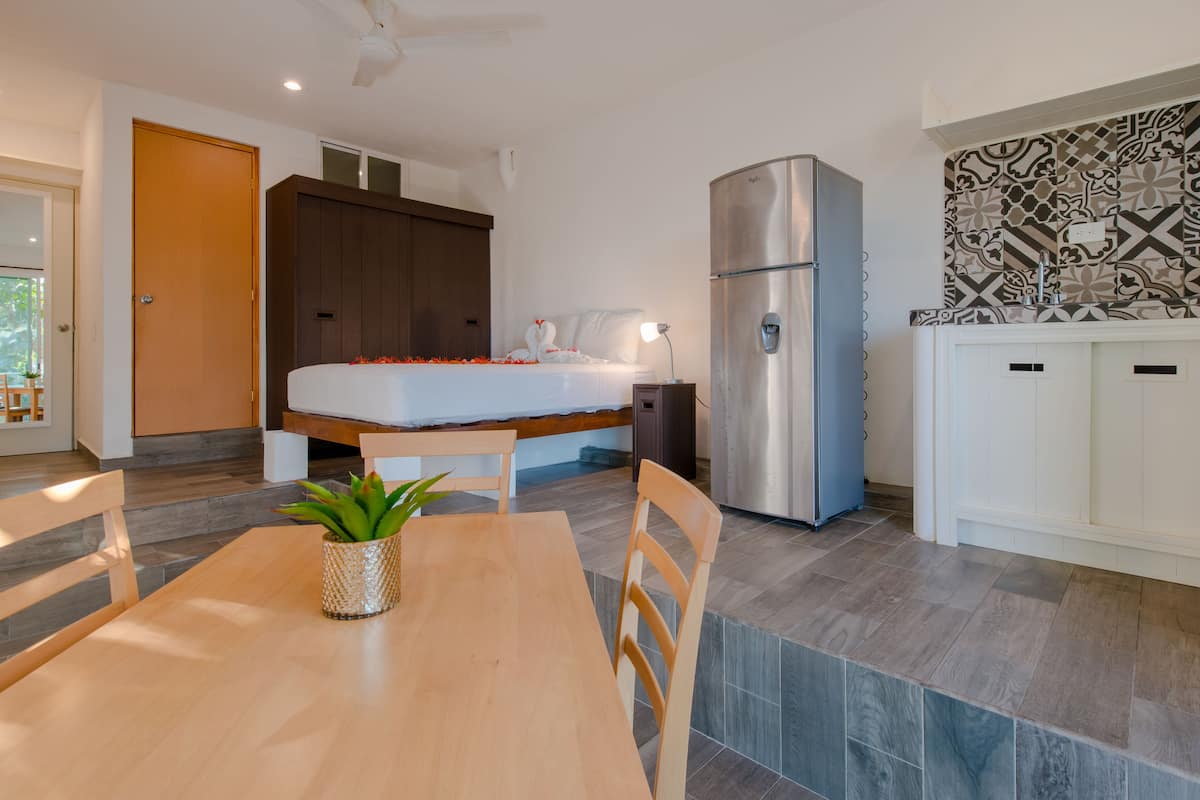 Un dormitorio moderno con cama, mesita de noche y un pequeño escritorio con lámpara. Hay un refrigerador y un armario blanco con azulejos estampados arriba.