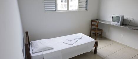 A small, clean room with a single bed, a desk with a computer, and a chair.