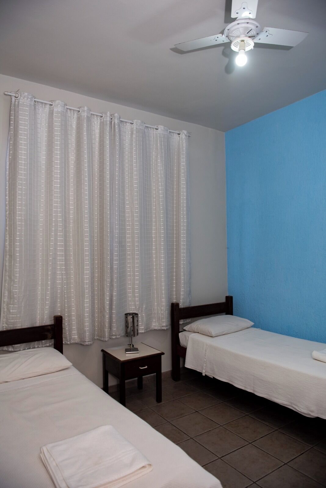 Una habitación con dos camas individuales, un ventilador de techo, una mesita con lámpara y una ventana con cortinas traslúcidas.