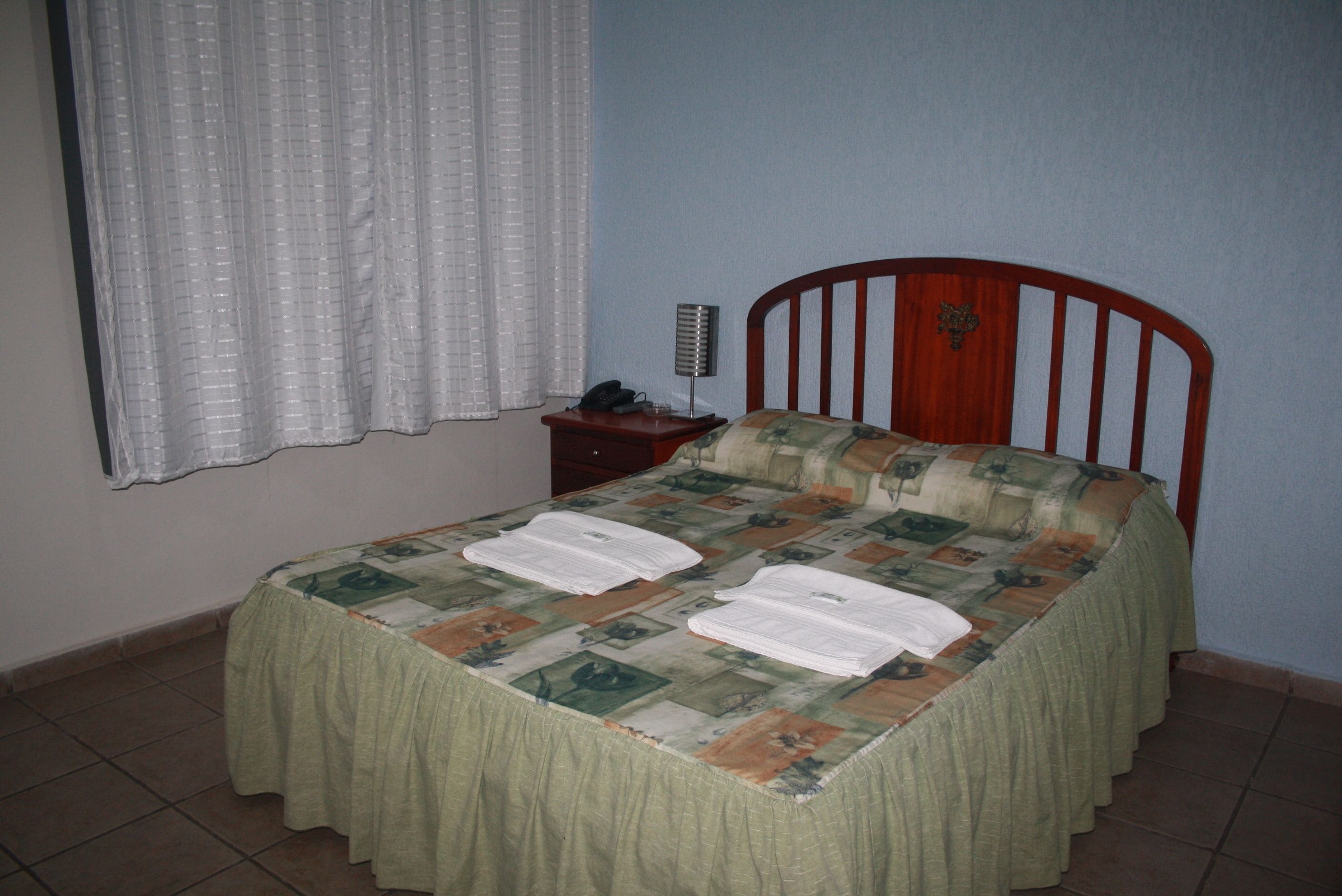 Un dormitori amb llit, una tauleta de nit, una làmpada i una finestra amb cortines.