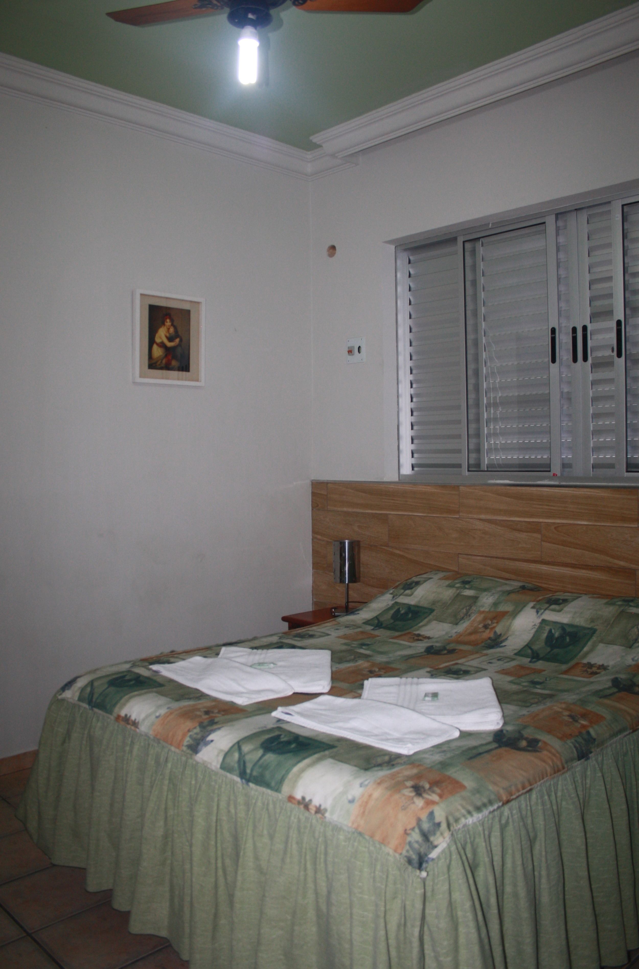 Ein Schlafzimmer mit einem Bett, einem Deckenventilator, einem Fenster mit Jalousien und einem kleinen Gemälde an der Wand.