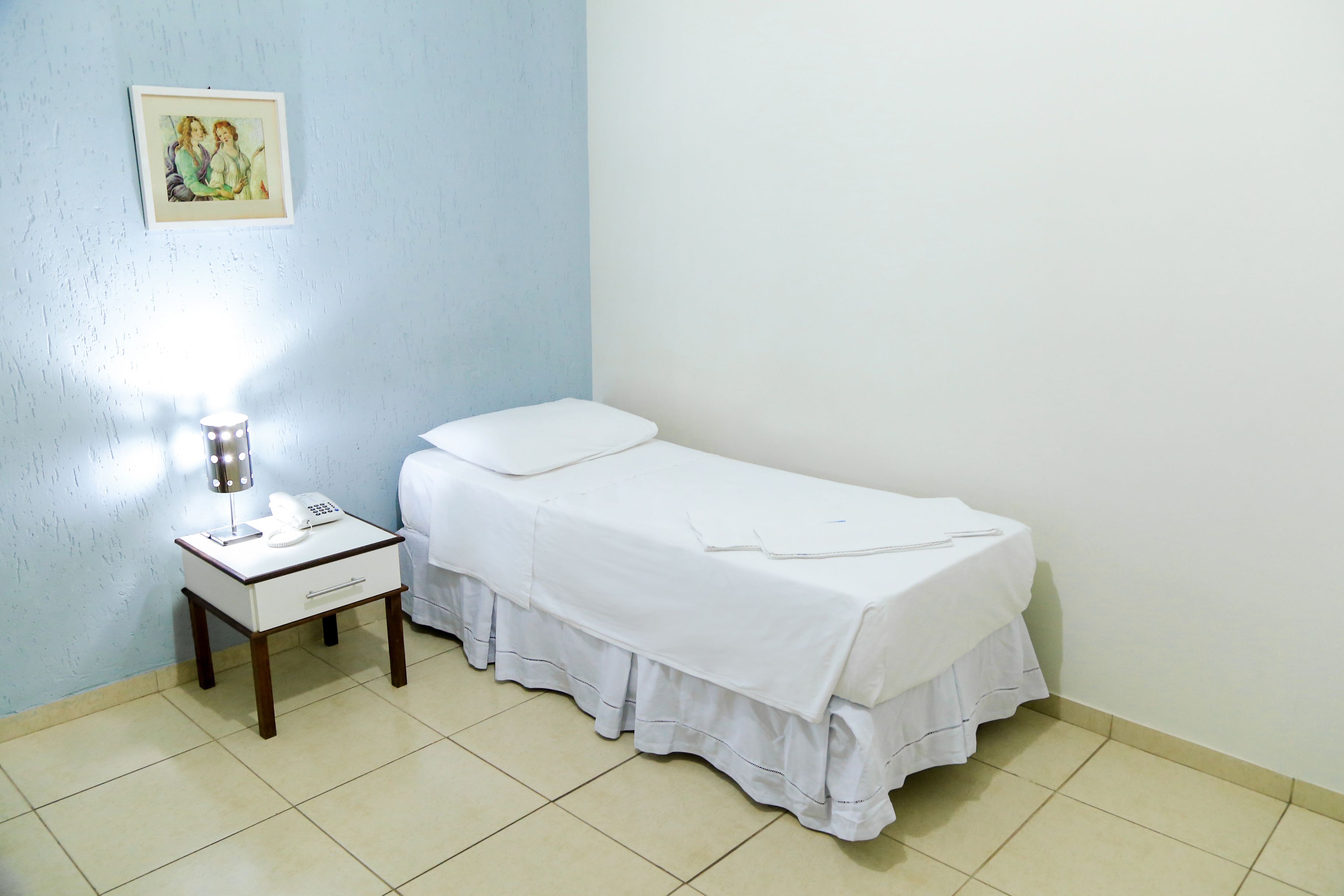 Una habitación pequeña y limpia con una cama individual, una mesita de noche con lámpara y un cuadro en la pared.