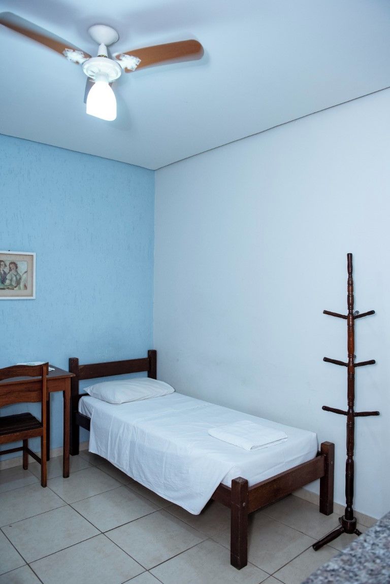 Una habitación pequeña con una cama individual, un escritorio con silla, un ventilador de techo y un perchero de madera.