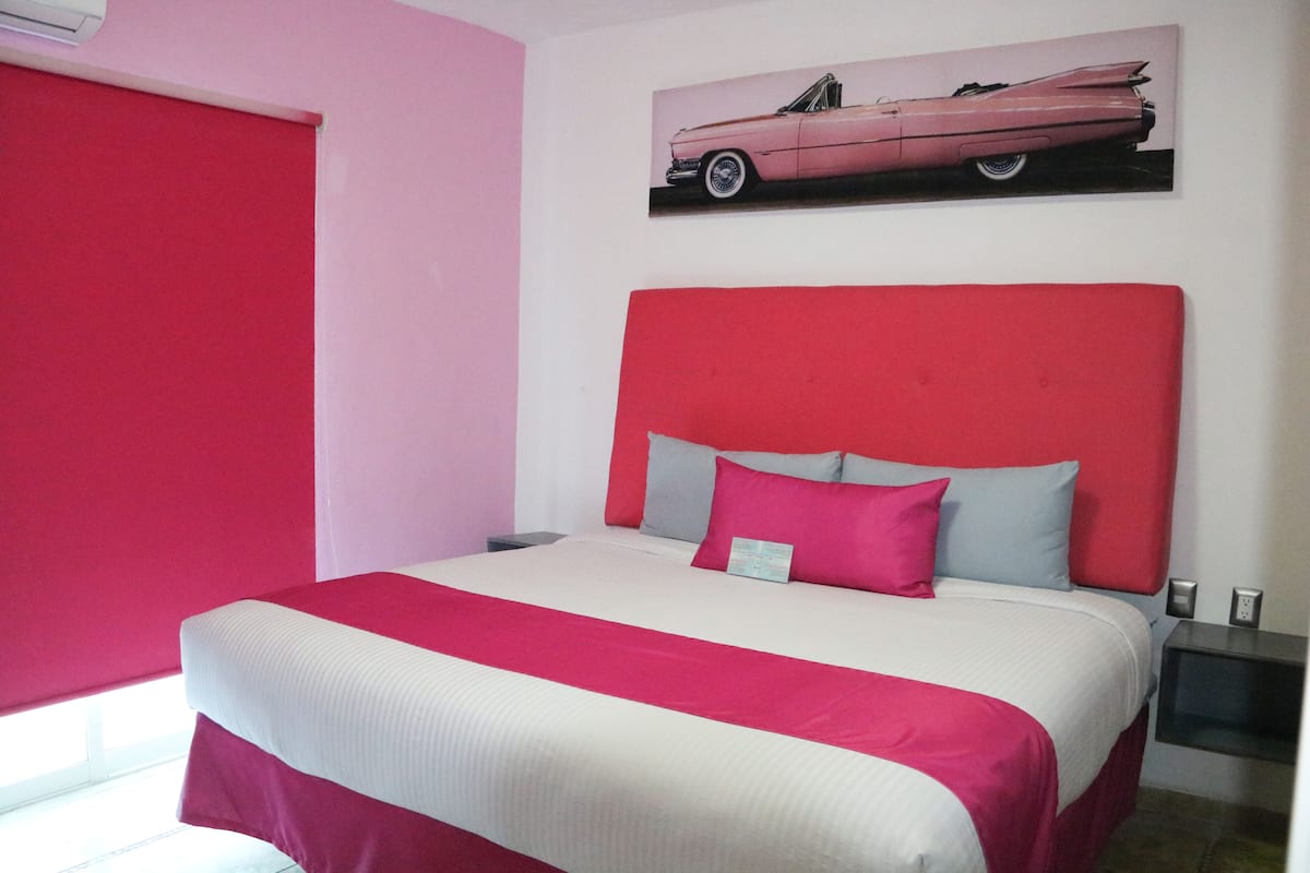 A bedroom with a bed, a headboard, a nightstand, a wall-mounted picture, and a window with blinds.