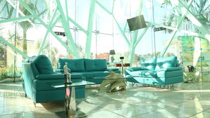 Un'area lounge moderna con divani color petrolio e tavolini in vetro, caratterizzata da un'ampia vetrata e una struttura in metallo.