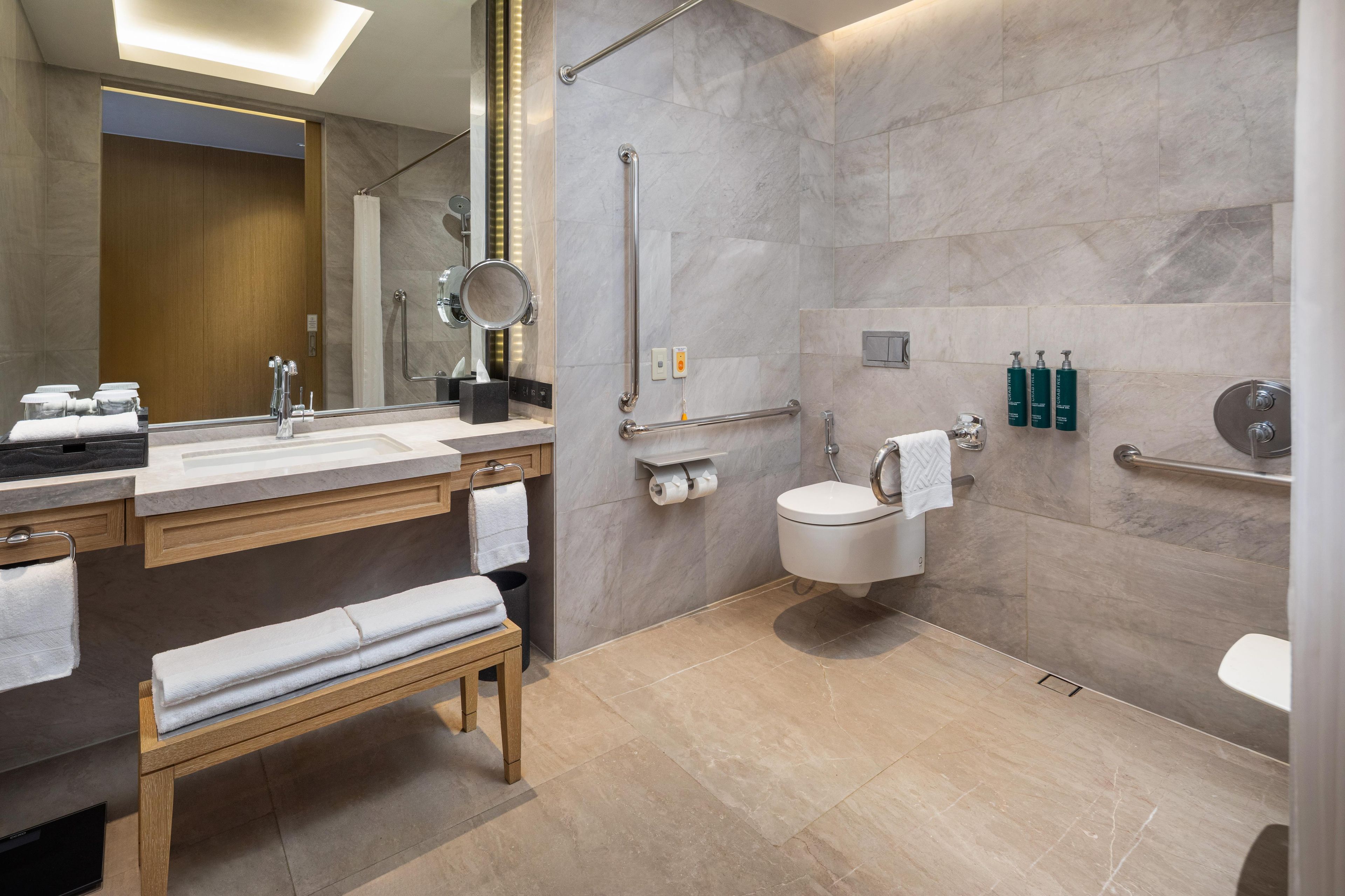 Un baño moderno con tocador, lavamanos, espejo, banco, inodoro y barras de apoyo.