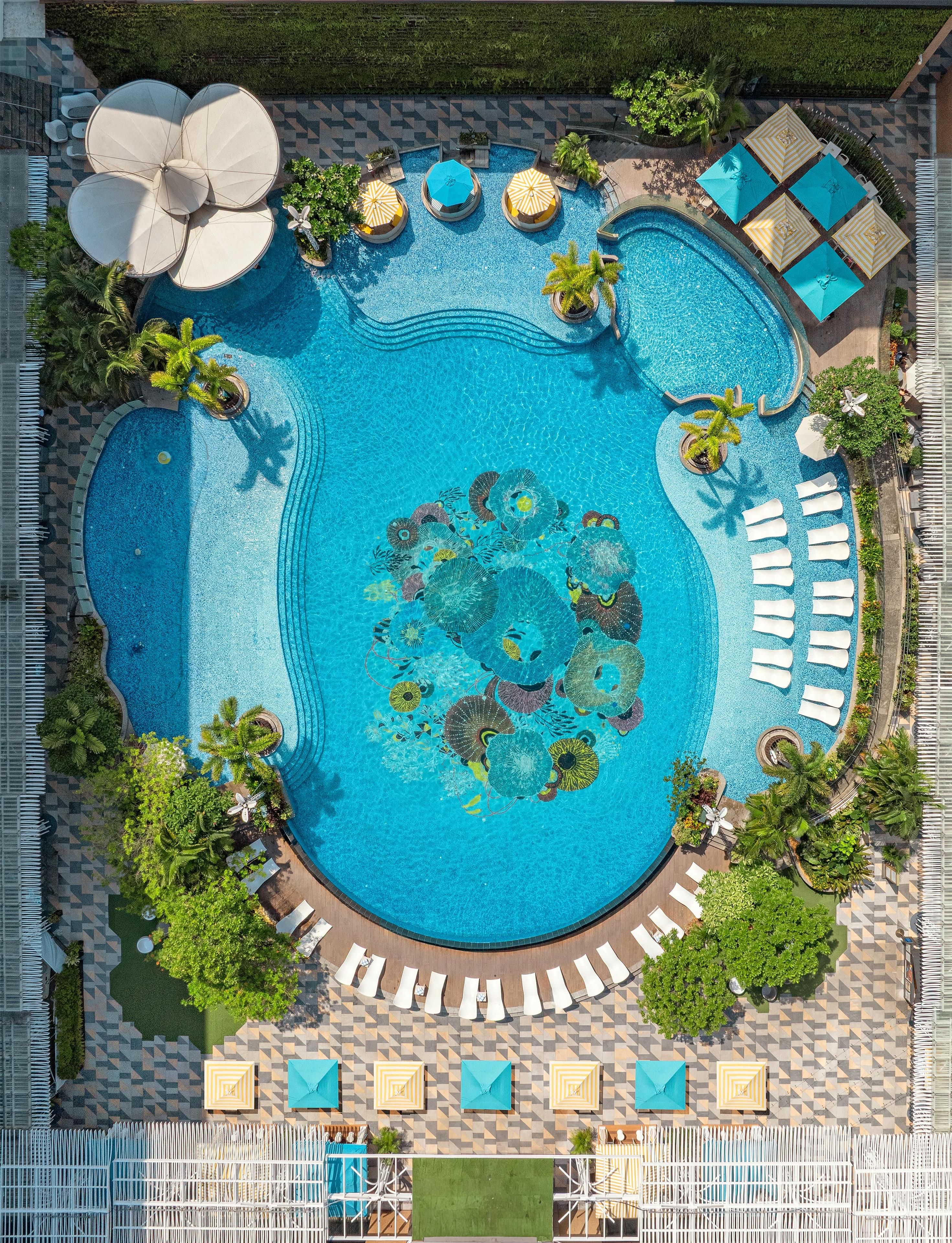 A large swimming pool with multiple sun loungers and umbrellas, surrounded by greenery and a tiled walkway.