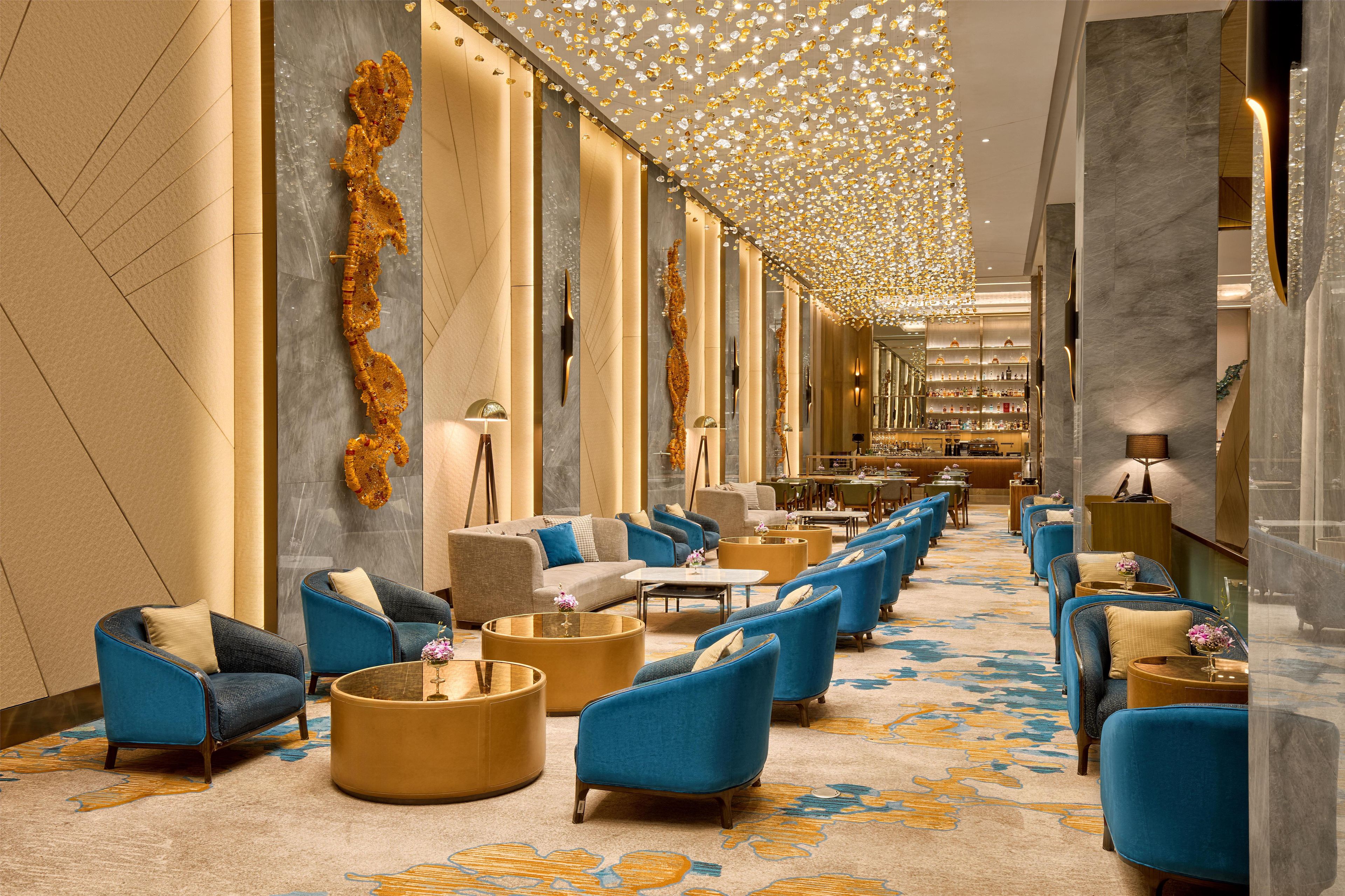 A hotel lobby with a long corridor, seating areas with blue armchairs, round coffee tables, and a patterned carpet.