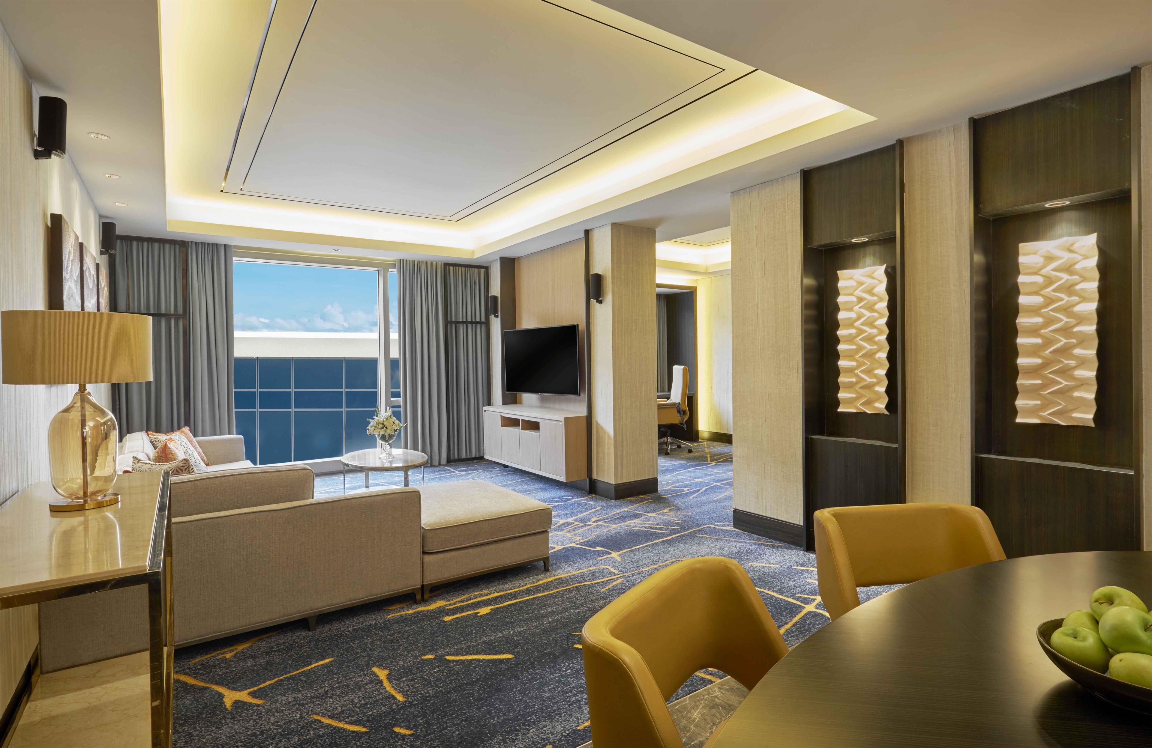Una habitación de hotel con sofá, una mesa de comedor con sillas, televisor y un ventanal con cortinas.