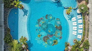 A large swimming pool with multiple sun loungers and umbrellas, surrounded by greenery and a tiled walkway.