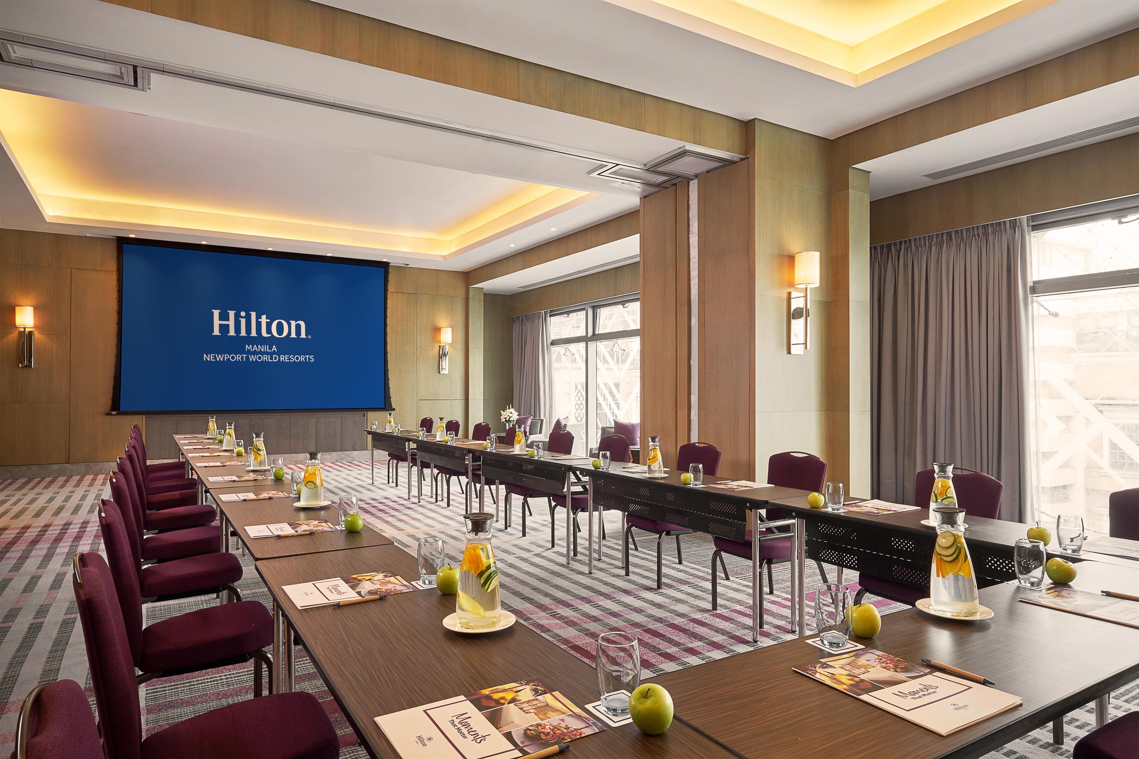 A conference room at the Hilton Manila Newport World Resorts with a long table set for a meeting, a large screen displaying the hotel's name, and a view of the city through the windows.
