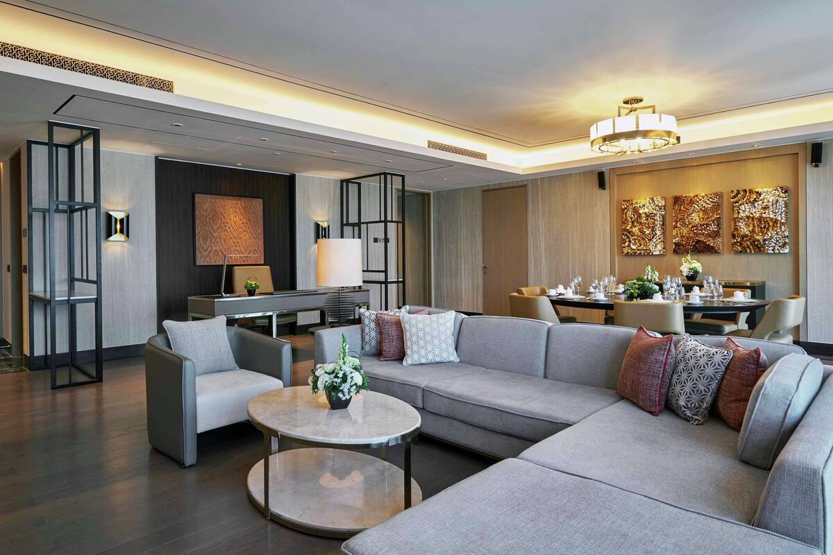A modern hotel lobby with a sectional sofa, a round coffee table, and a dining area with a chandelier.