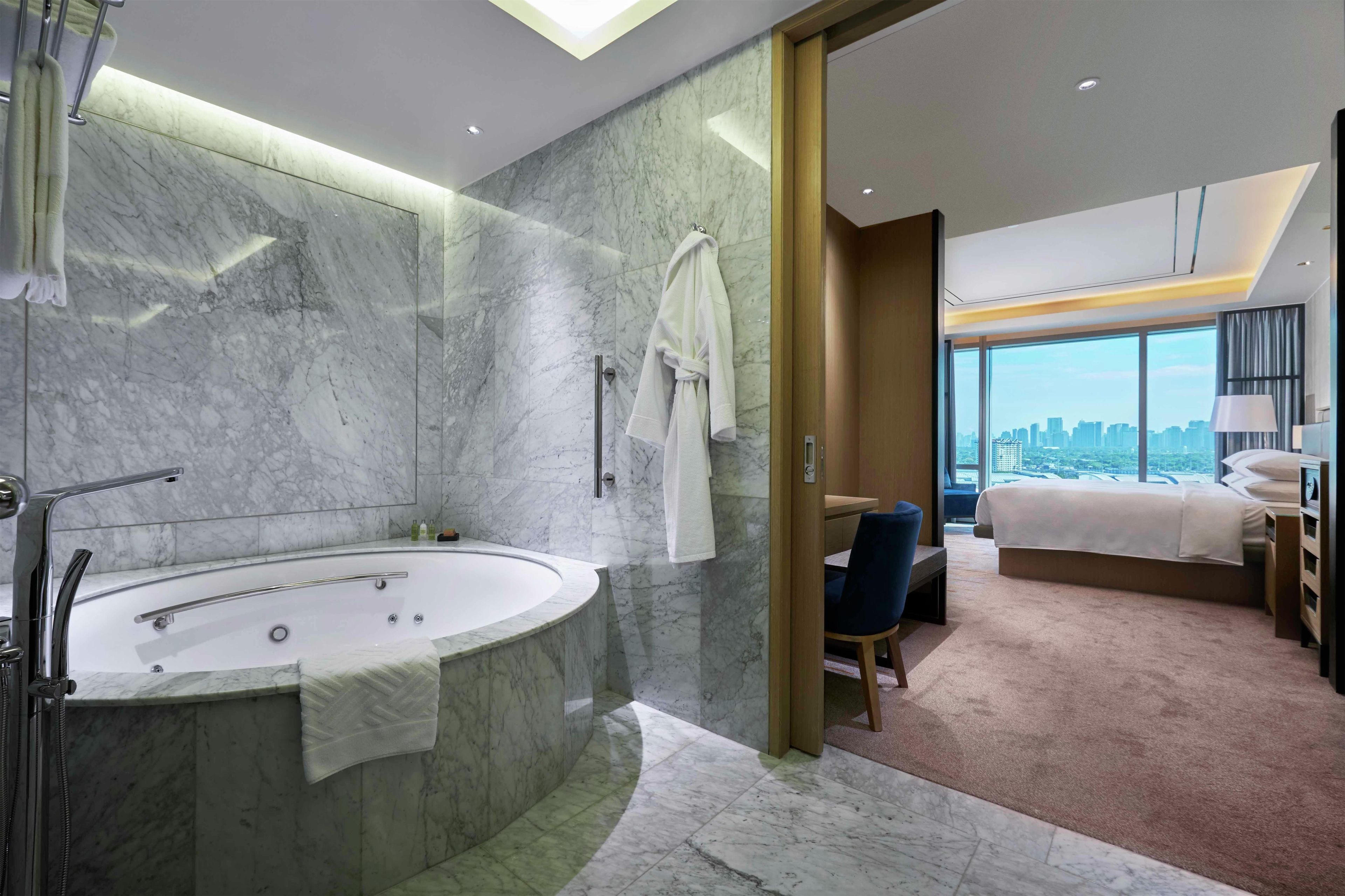 A modern hotel room with a marble bathroom, a large bathtub, and a city view.