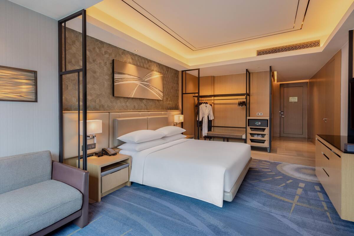 A modern hotel room with a large bed, a seating area, a wardrobe, and a desk with a lamp.
