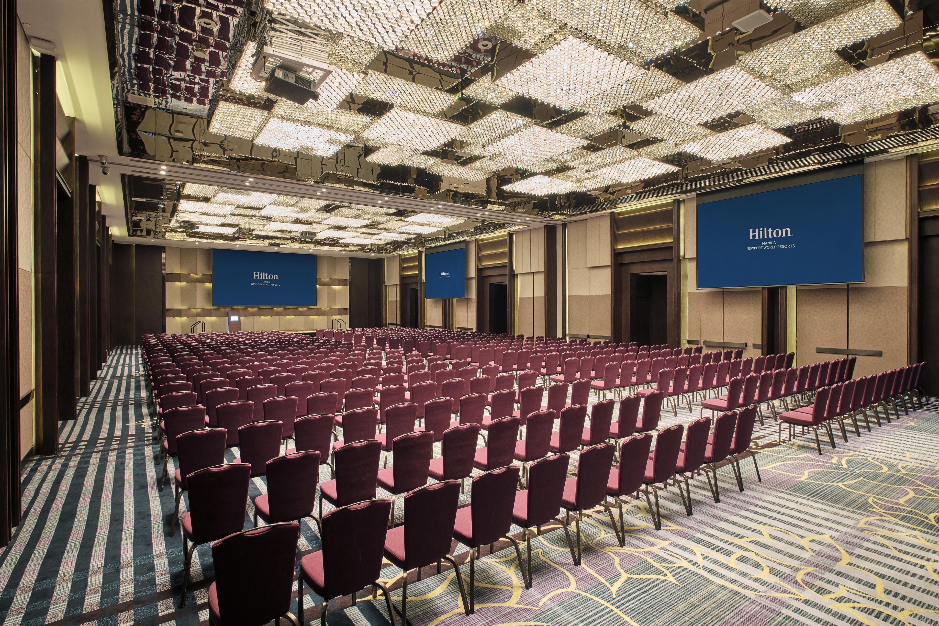 Un salón de conferencias amplio con filas de sillas rojas, alfombra estampada y un techo con una cuadrícula de luces.