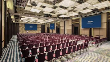 Un salón de conferencias amplio con filas de sillas rojas, alfombra estampada y un techo con una cuadrícula de luces.