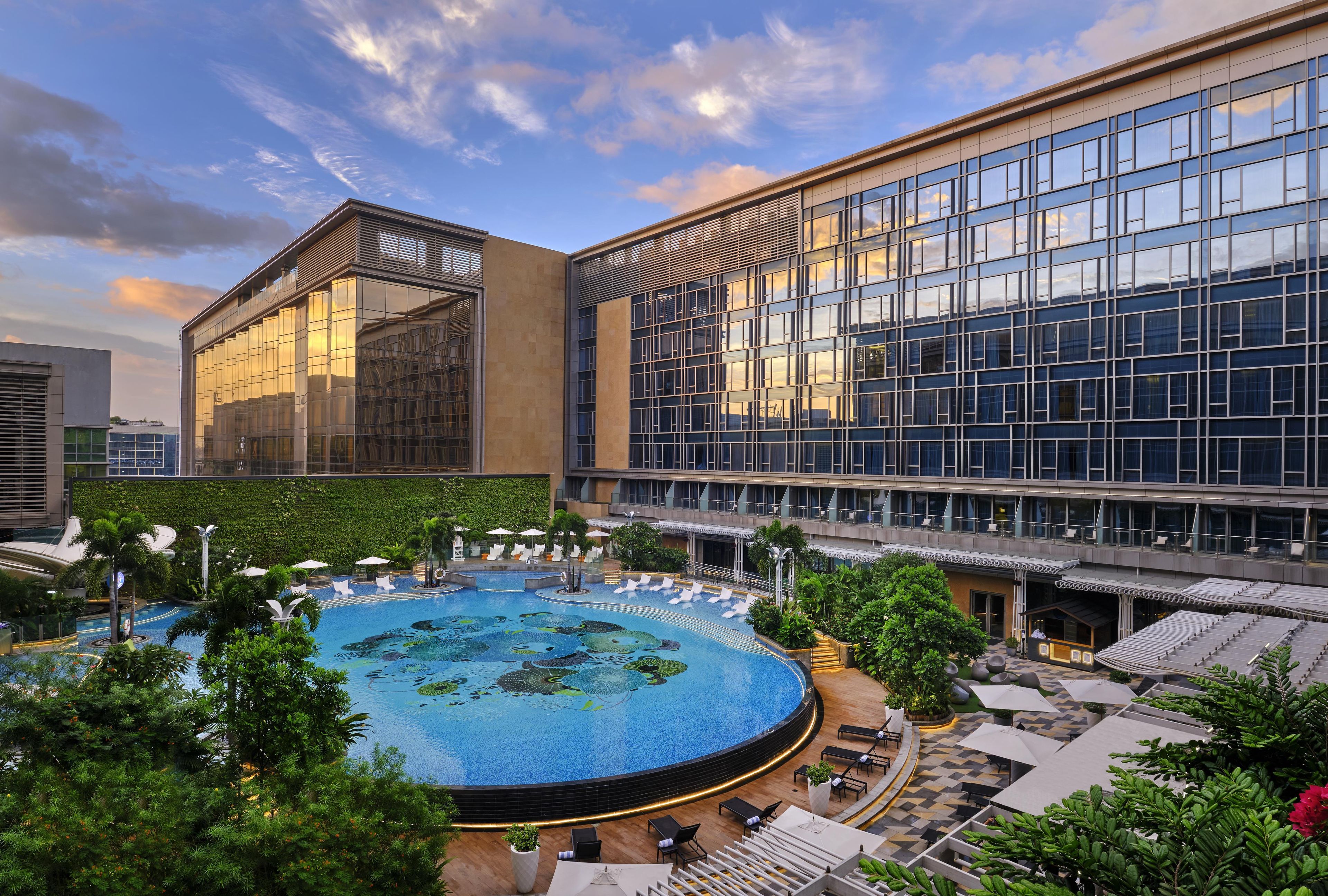 A modern hotel with a large swimming pool surrounded by lounge areas and greenery.