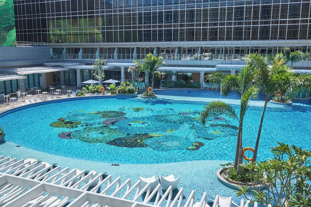 A hotel pool area with a clear blue pool, lounge chairs, and palm trees.