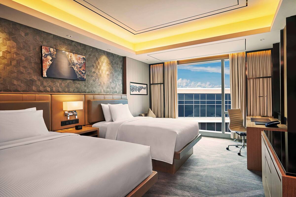 A hotel room with two beds, a desk, a chair, a TV, and a large window with curtains.