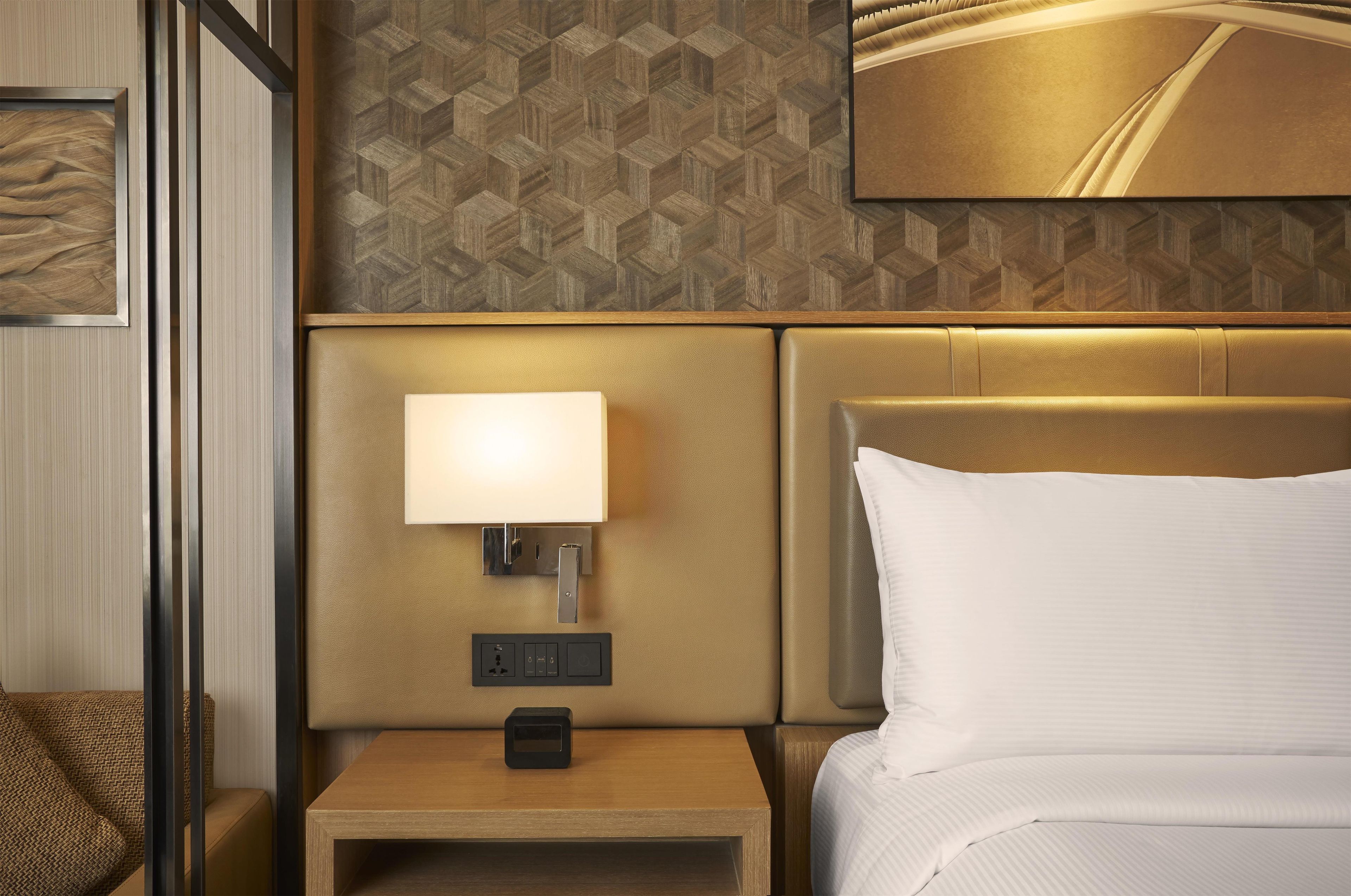 Una habitación de hotel con una cama, una mesita de noche con lámpara y un panel de control en la pared.