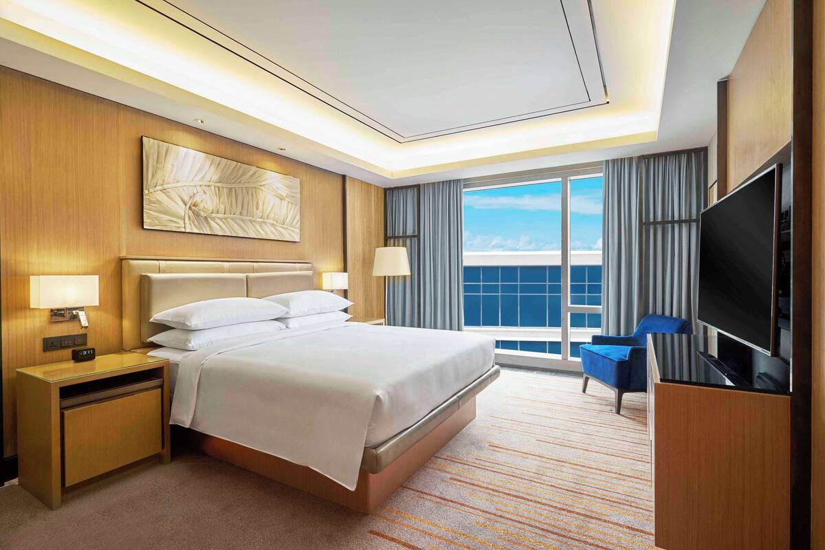 A hotel room with a large bed, a TV, a desk with a lamp, and a view of the sky through the window.
