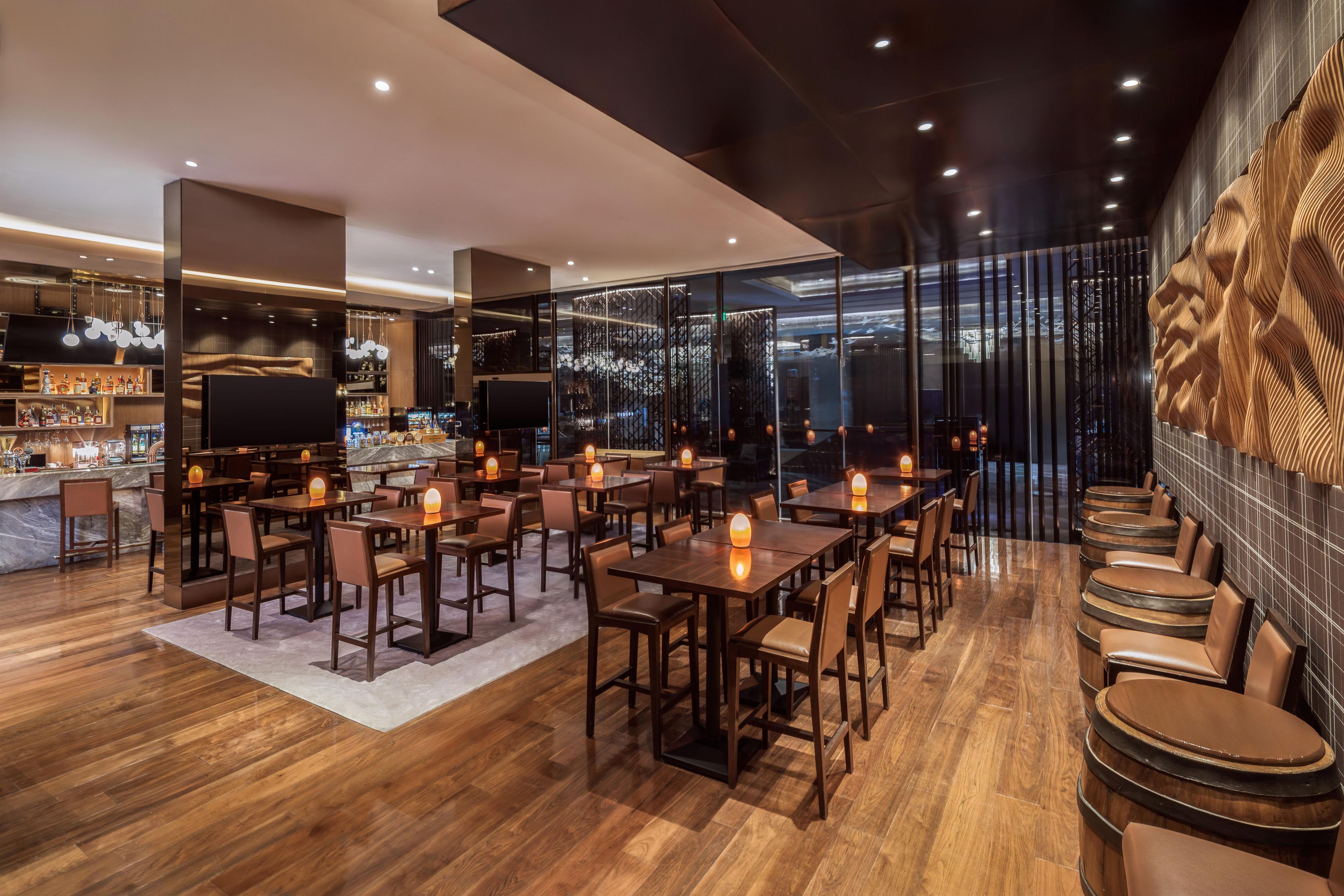 A modern restaurant with wooden flooring, tables with candle-like lighting, and a bar area with shelves of bottles.