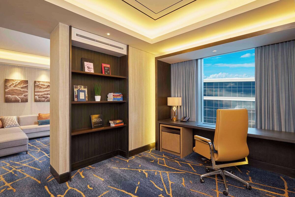 A hotel room with a sofa, a desk with a chair, a bookshelf, and a large window with curtains.