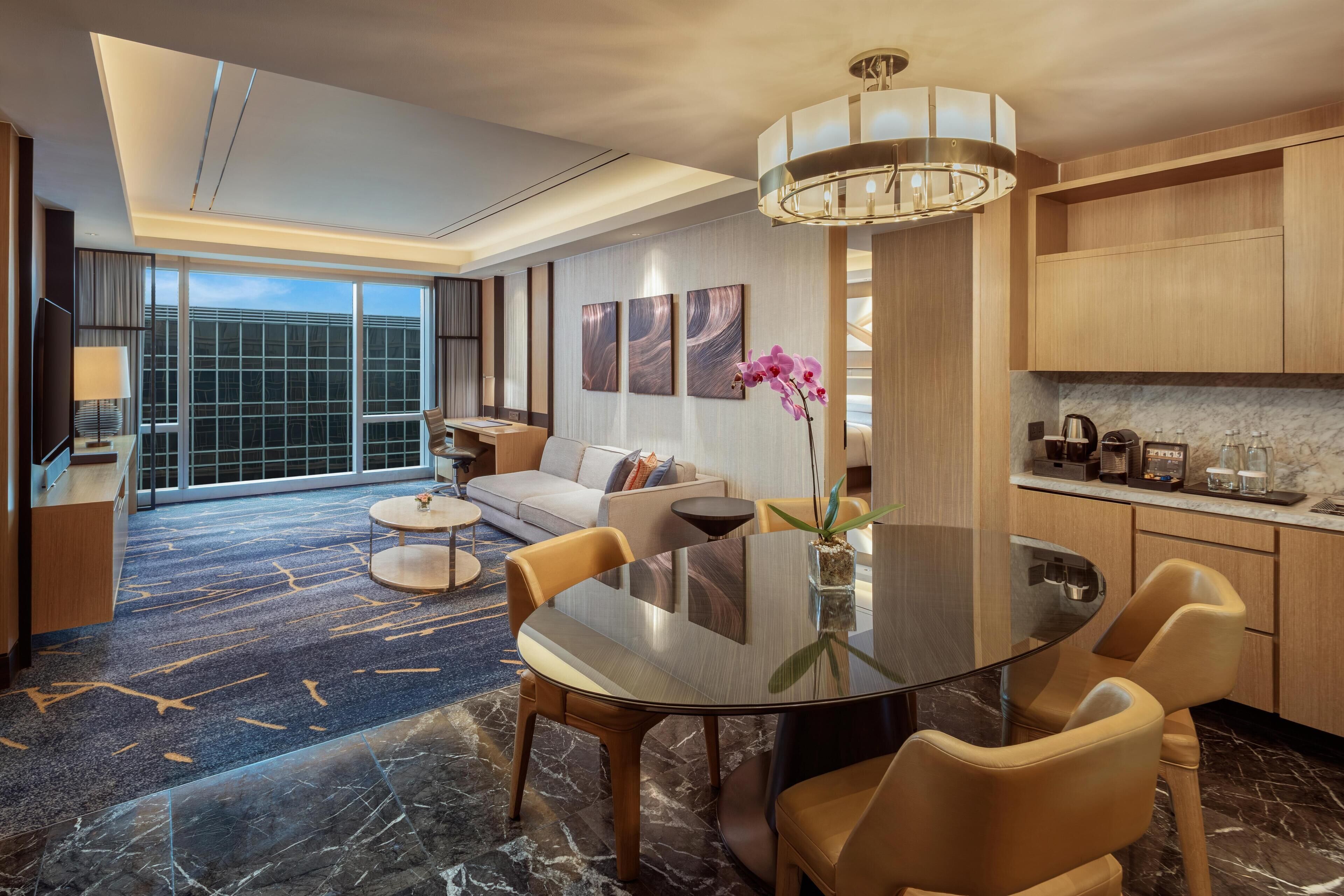 A modern hotel room with a large dining table, comfortable seating, a sofa, a TV, and a large window offering a city view.