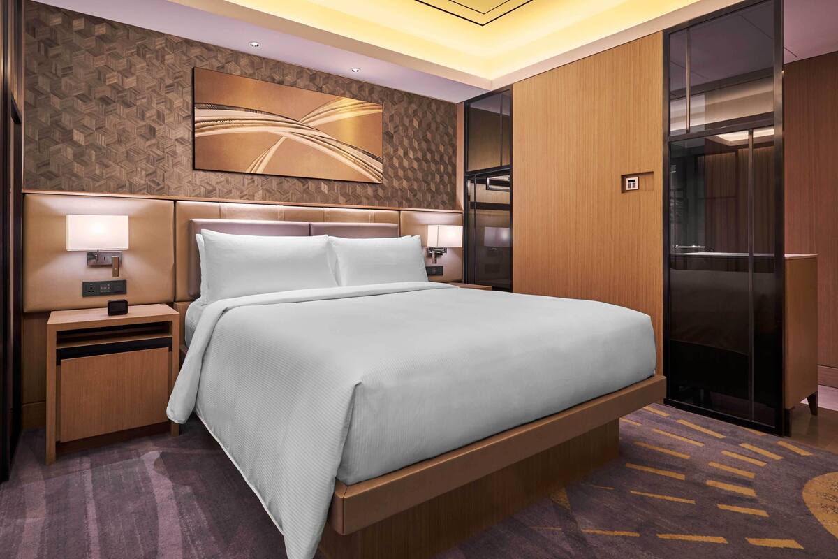 A modern hotel room with a large bed, wooden headboard, two bedside tables with lamps, a wall-mounted artwork, and a sliding door closet.