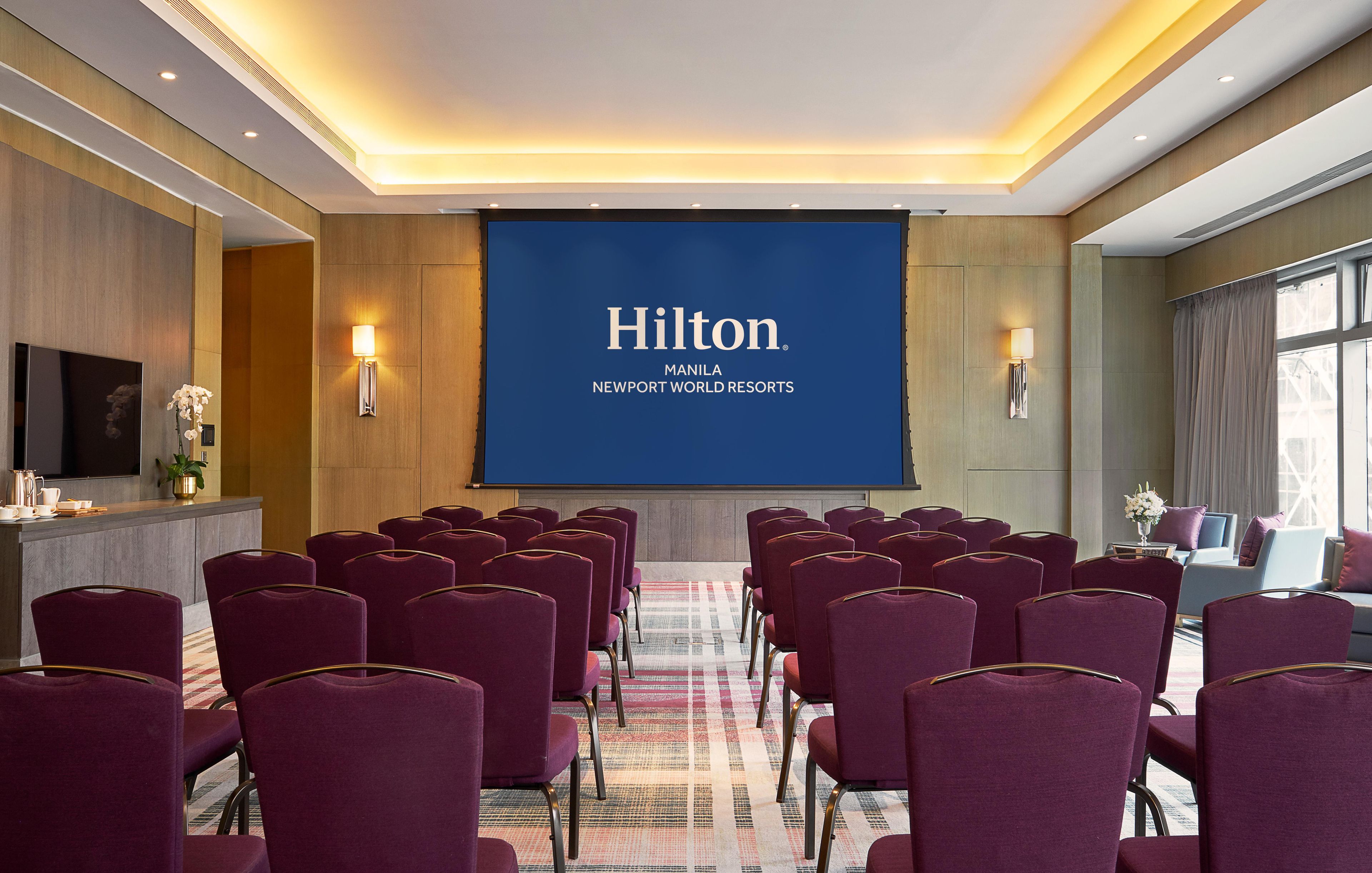A conference room with rows of purple chairs, a large screen displaying 'Hilton Manila Newport World Resorts', and a wooden panel wall.