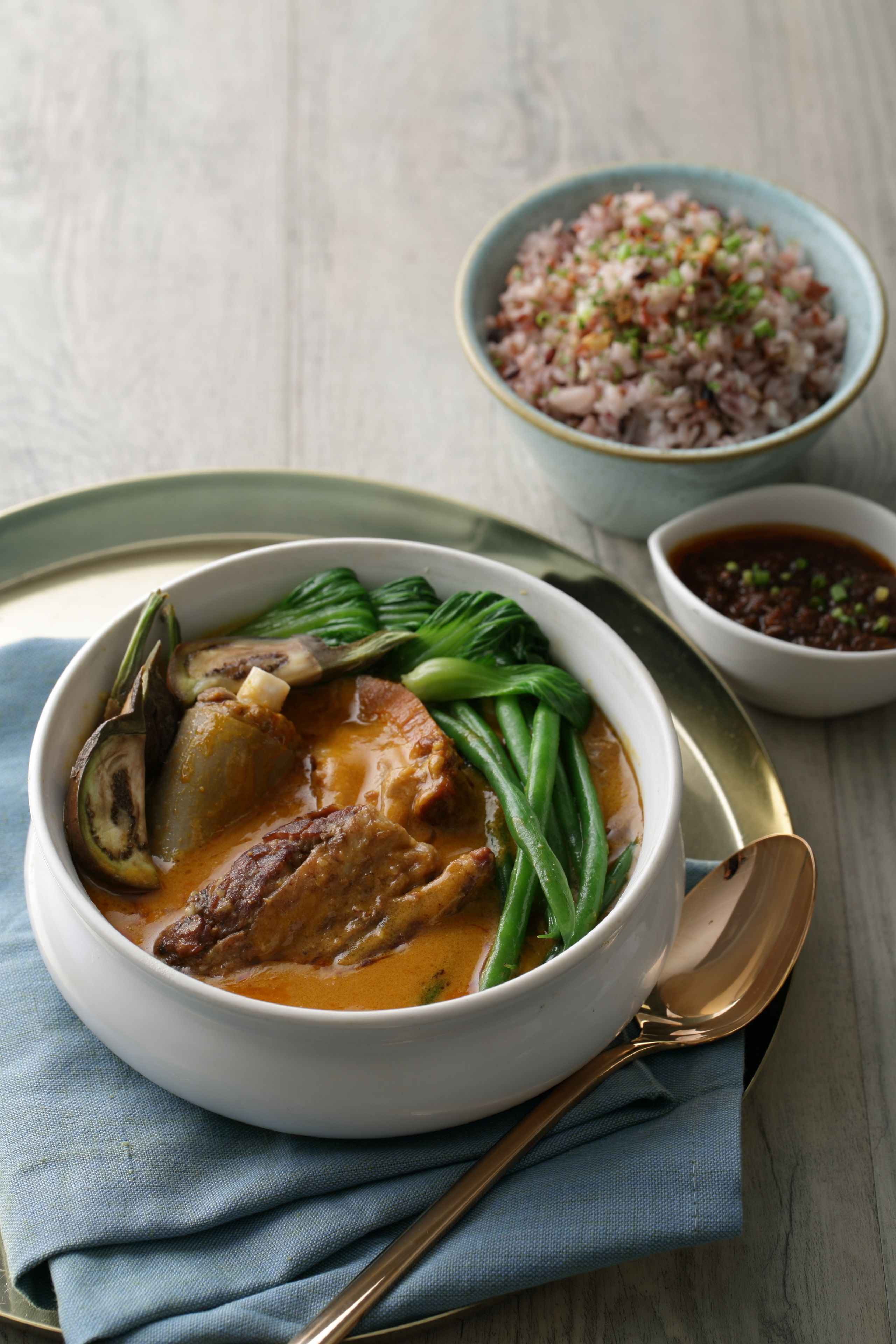 A bowl of soup with vegetables and meat, a spoon, and a small bowl of sauce on a table.