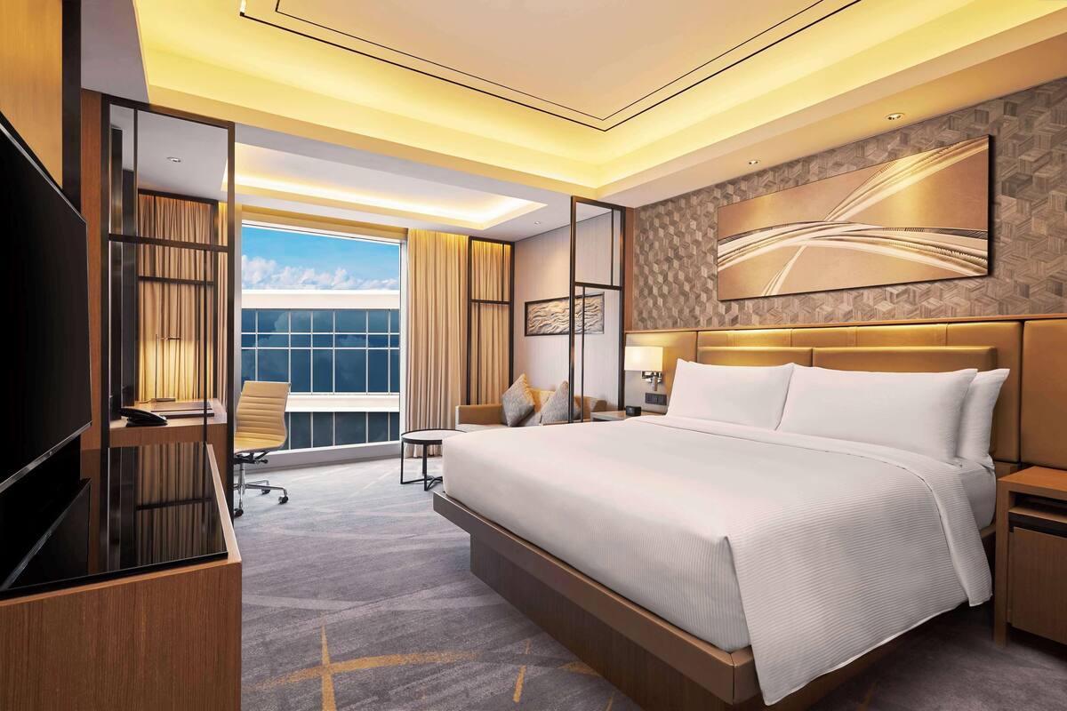 A modern hotel room with a large bed, a desk, a chair, and a view of the sky through a large window.