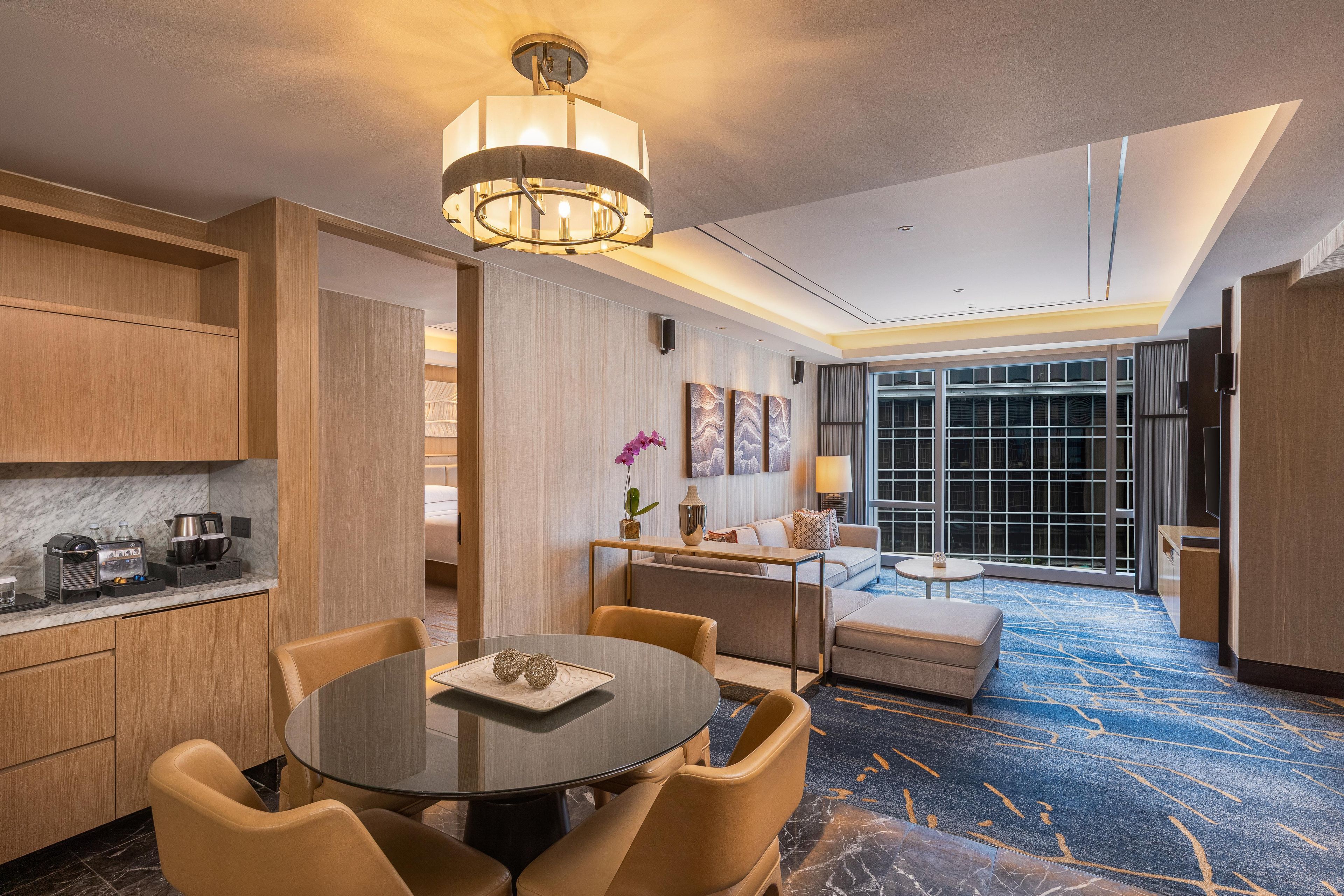 Quarto de hotel moderno com área de refeições, sofá, mesa pequena e uma janela grande.