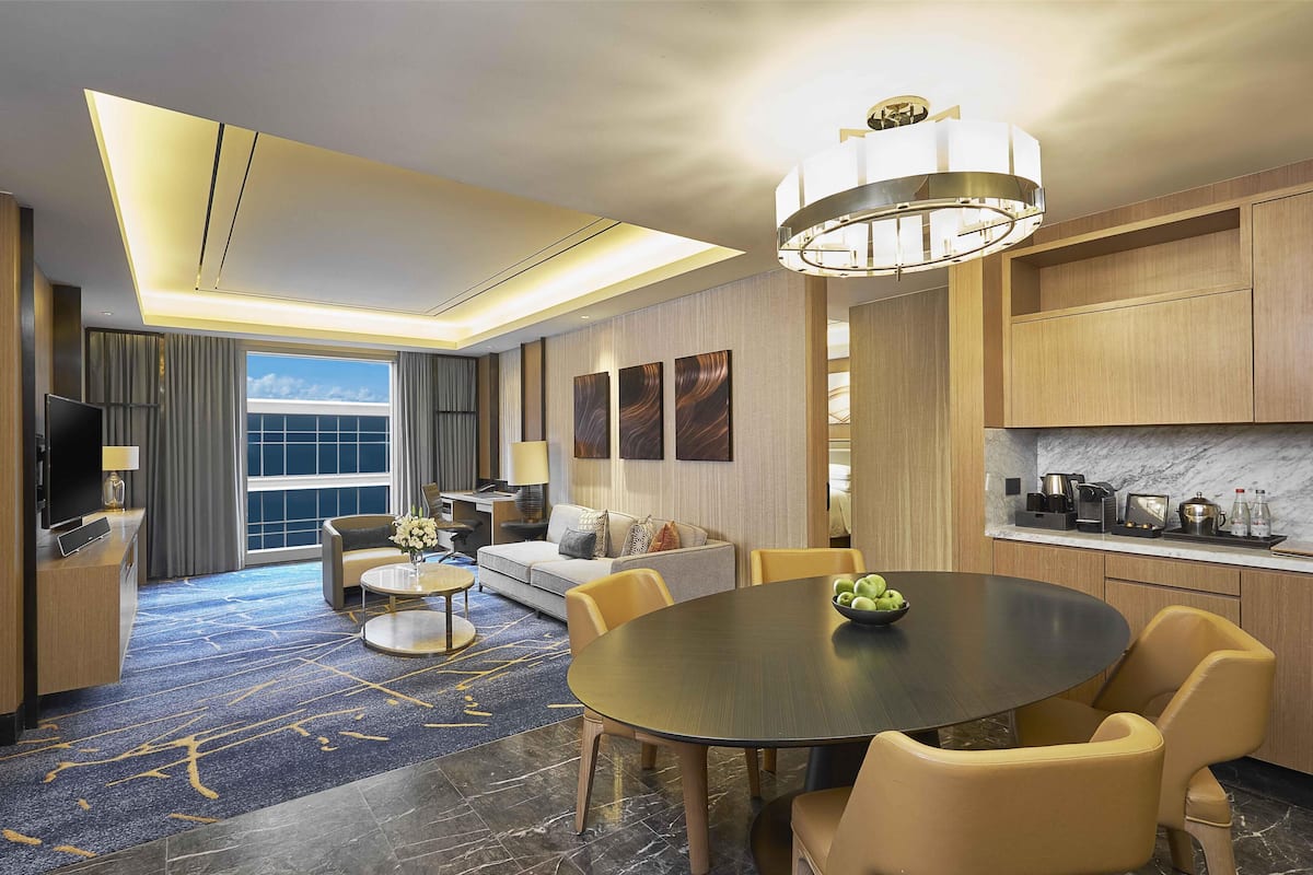 A modern hotel room with a dining area, a sofa, a TV, and a large window.
